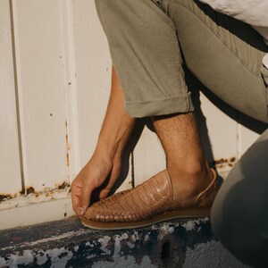 May include: Tan leather woven shoe with a rubber sole. The shoe has a slip-on design and a textured upper. The shoe is worn with olive green pants and a white shirt.