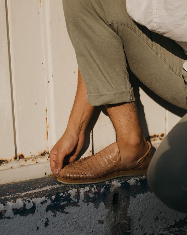 May include: Tan leather woven shoe with a rubber sole. The shoe has a slip-on design and a textured upper. The shoe is worn with olive green pants and a white shirt.