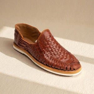 May include: A brown leather woven shoe with a white sole. The shoe has a low profile and a slip-on design. The woven pattern covers the top of the shoe.