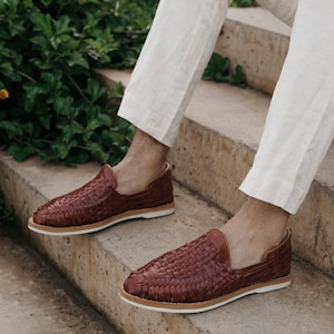 May include: A pair of brown leather woven loafers with white soles. The shoes are displayed on a concrete staircase, showcasing their intricate design and casual style. The loafers are a rich, warm brown color.