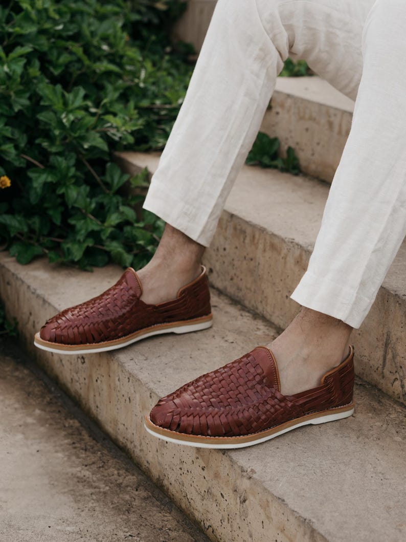 May include: A pair of brown leather woven loafers with white soles. The shoes are displayed on a concrete staircase, showcasing their intricate design and casual style. The loafers are a rich, warm brown color.