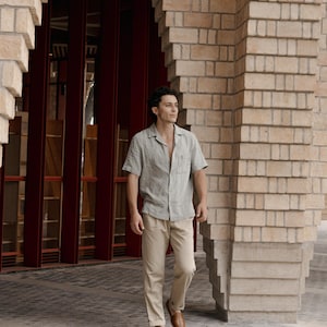 May include: A man wearing a light green short-sleeved button-down shirt and tan pants. He is wearing brown woven leather shoes. The background features a brick building with red doors.