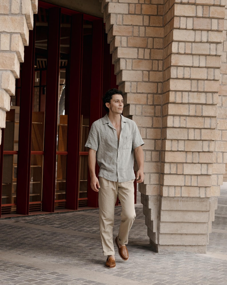 May include: A man wearing a light green short-sleeved button-down shirt and tan pants. He is wearing brown woven leather shoes. The background features a brick building with red doors.