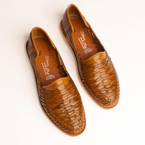 May include: A pair of brown leather woven huarache shoes. The shoes have a rounded toe and a low profile. The soles are white with a stitched edge. The interior of the shoe has text that reads "Mexico en la Piel".