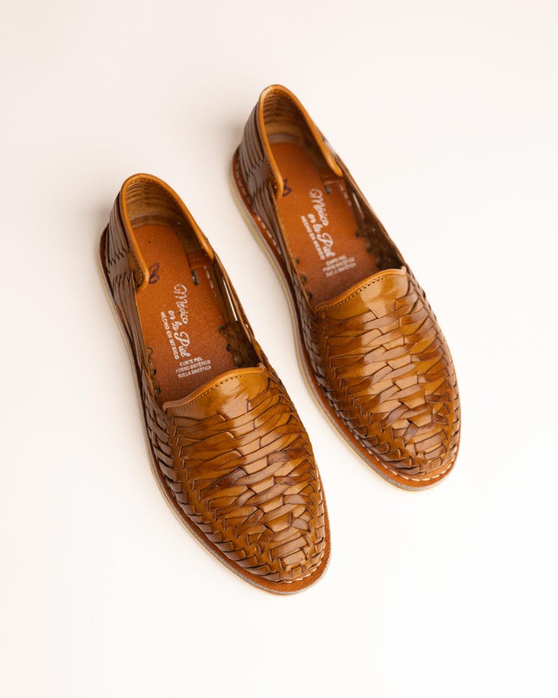 May include: A pair of brown leather woven huarache shoes. The shoes have a rounded toe and a low profile. The soles are white with a stitched edge. The interior of the shoe has text that reads "Mexico en la Piel".