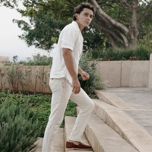 May include: A person wearing a white short-sleeve shirt and light-colored pants. The person is wearing brown woven leather loafers. The person is walking up a set of concrete stairs in an outdoor setting with greenery.
