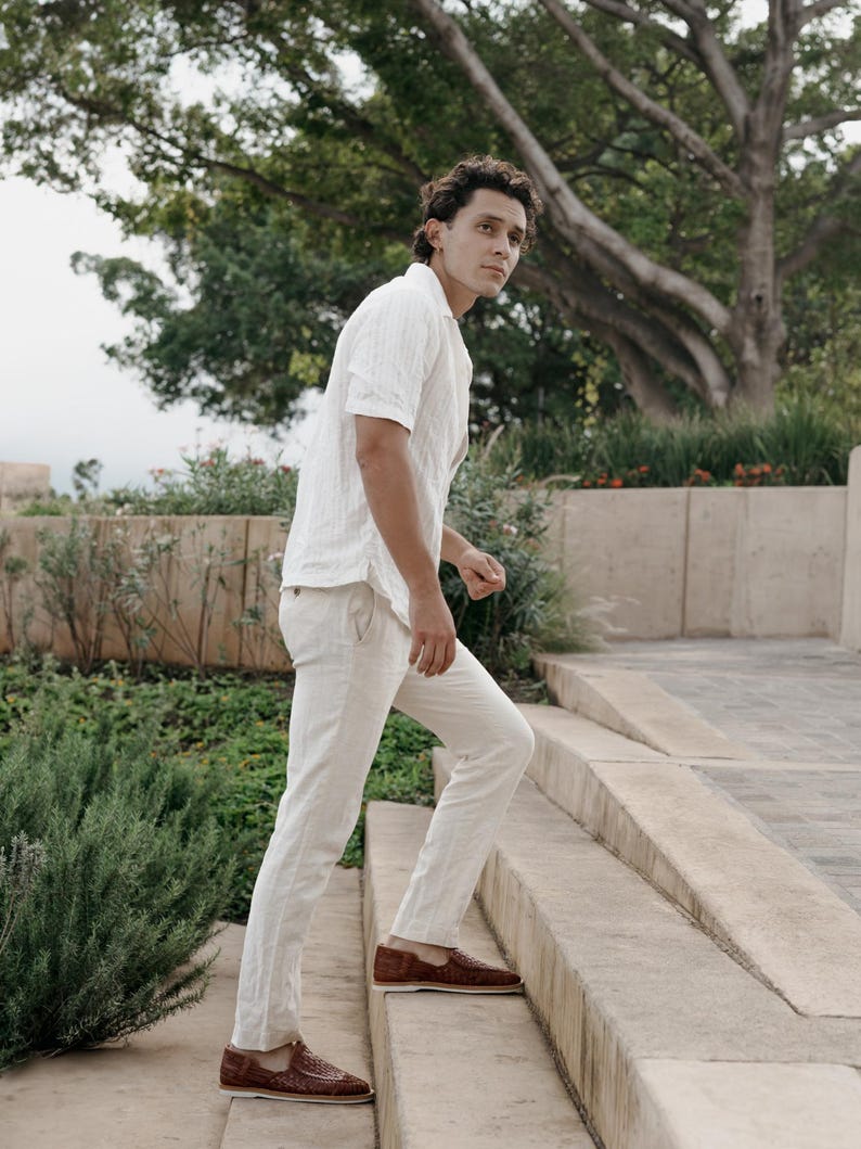 May include: A person wearing a white short-sleeve shirt and light-colored pants. The person is wearing brown woven leather loafers. The person is walking up a set of concrete stairs in an outdoor setting with greenery.