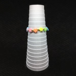 May include: A white plastic ring sizer with a rainbow star-shaped beaded ring on the size 6.75 ring. The sizer has markings from size 5.25 to size 9.