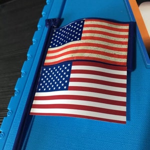 May include: A small American flag with a blue pole, the flag is printed on a white background with red and white stripes and white stars on a blue field. The flag is attached to a blue plastic pole.