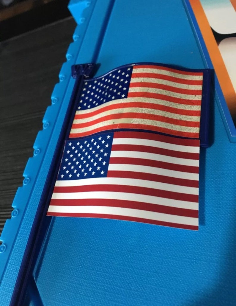 May include: A small American flag with a blue pole, the flag is printed on a white background with red and white stripes and white stars on a blue field. The flag is attached to a blue plastic pole.