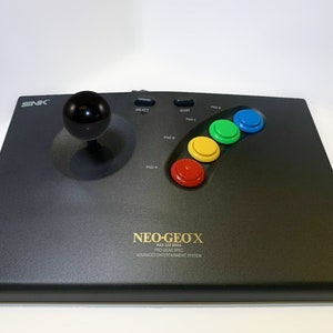 May include: A black arcade joystick controller featuring a spherical black handle, two small oval buttons labeled Select and Start, and four circular colored buttons in red, yellow, green, and blue. The console is branded with SNK and NEO GEO X.