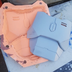 May include: A light gray plastic mold with a detailed design. The mold is used to create a 3D printed object. The object is a light gray plastic piece with a geometric design.