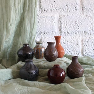 May include: A collection of seven small ceramic vases in various shapes and earth-toned glazes, including dark brown, reddish-brown, and mottled grey, arranged in a cluster with narrow necks and rounded bodies on a draped fabric surface.