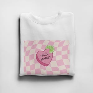 May include: White t-shirt with a pink and white checkered pattern and a pink heart with the text "Spicy Margs" on it. A green lime wedge is on top of the heart.