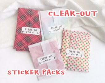 Puede incluir: Cuatro paquetes de pegatinas envueltos en papel con patrones coloridos. Los paquetes están etiquetados con "CLEAR-OUT *Pack!!" y "CLEAR-OUT ★° packs Sr". El texto "STICKER PACKS" está escrito debajo de los paquetes.