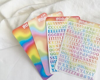 Puede incluir: Cuatro hojas de pegatinas de color arcoíris con el texto "Lots Liv. stef" y "Lots" impreso en ellas. Las pegatinas presentan un patrón de tie-dye arcoíris y letras del alfabeto en un degradado arcoíris.
