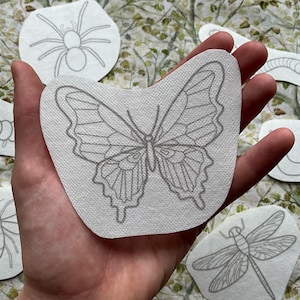 Puede incluir: Un conjunto de parches para planchar con diseños de varios insectos, incluyendo una mariposa, una araña, una oruga, un caracol, una libélula y una abeille. Los parches son blancos con contornos negros.
