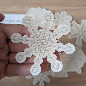 May include: A collection of white snowflake-shaped patches with gray embroidered details. The patches vary in size and design, with a prominent snowflake in the center. The patches are on a light-colored surface, suggesting a winter or holiday theme.
