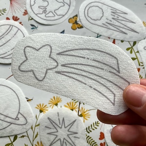 May include: A selection of white, die-cut stickers with space-themed illustrations. Designs include a planet with rings, a starburst, a comet, a constellation, and the Earth. The stickers are held by a hand against a floral background.