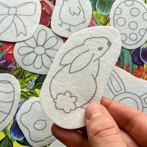 Könnte beinhalten: Ein Set mit 10 weißen Filz-Ostereiershapen mit verschiedenen Designs, darunter ein Hase, eine Blume, eine Schleife, ein Küken und ein Ei. Die Designs sind in Grau umrandet.