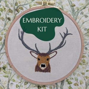 Könnte beinhalten: Ein Stickset mit einem Hirschmotiv. Der Hirsch ist in Braun-, Beige- und Grautönen mit detailliertem Geweih gestickt. Der Text "EMBROIDERY KIT" wird auf einer grünen Form angezeigt. Das Set befindet sich in einem Holzreifen.