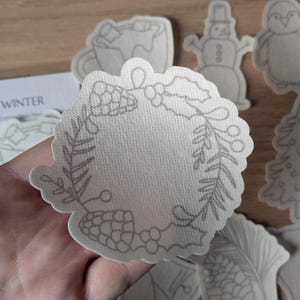 May include: A hand holds a light-coloured, die-cut wreath with a detailed grey outline. Other winter-themed die-cuts, including a snowman, penguin, and Christmas tree, are visible. The words "COSY WINTER" are partially visible in the background.