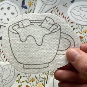 Könnte beinhalten: Eine weiße Filzapplikation einer Tasse mit heißer Schokolade und Marshmallows. Die Tasse ist mit schwarzem Garn umrandet und hat einen Henkel.
