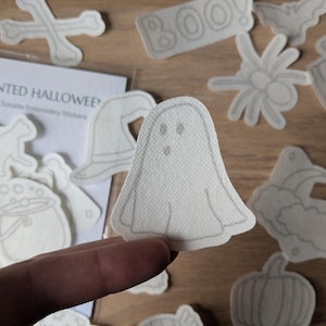 Könnte beinhalten: Eine Sammlung wasserlöslicher Stickaufkleber mit Halloween-Motiven. Die Aufkleber zeigen einen Geist, einen Hexenhut, eine Spinne, einen Kürbis und das Wort "BOO!". Die Aufkleber sind weiß mit grauen Umrissen und liegen auf Holz.