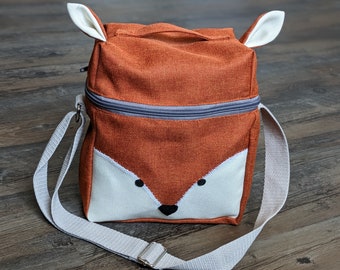 Könnte beinhalten: Eine braune Canvas-Lunch-Tasche mit einem Fuchsgesicht auf der Vorderseite. Die Tasche hat einen weißen Trageriemen und einen Reißverschluss.