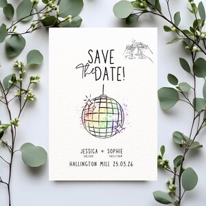 May include: A white wedding invitation card featuring the text "SAVE the DATE!" and a rainbow disco ball illustration. The names "JESSICA + SOPHIE" and the date "25.03.26" are printed on the card. The card is surrounded by green foliage.