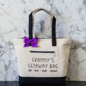 May include: A white canvas tote bag with black handles and a black bottom. The bag has a zippered pocket and the text "Grammy's Getaway Bag visit - love - spoil - repeat" printed on the front.