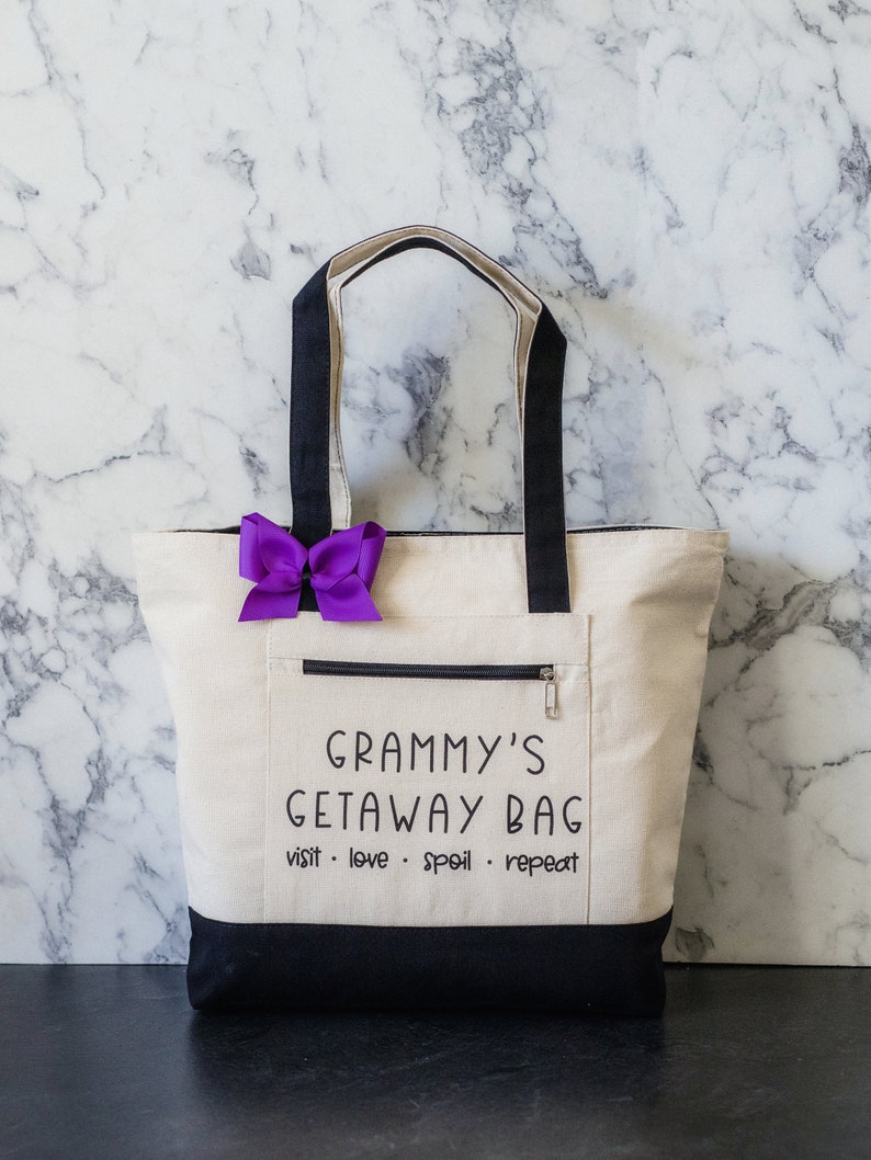 May include: A white canvas tote bag with black handles and a black bottom. The bag has a zippered pocket and the text "Grammy's Getaway Bag visit - love - spoil - repeat" printed on the front.
