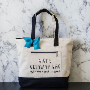 May include: A white canvas tote bag with black handles and a black bottom. The bag has a zippered pocket on the front and the text "Gigi's Getaway Bag visit. love. spoil. repeat" printed in black.