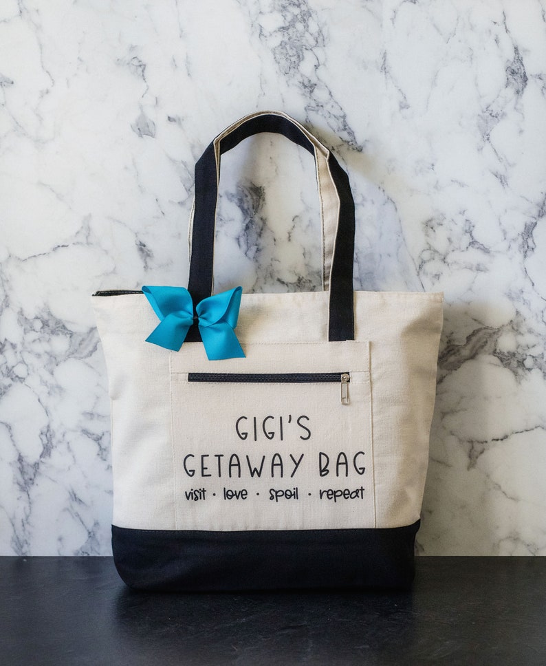 May include: A white canvas tote bag with black handles and a black bottom. The bag has a zippered pocket on the front and the text "Gigi's Getaway Bag visit. love. spoil. repeat" printed in black.