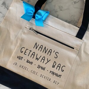 May include: A white canvas tote bag with black handles and a black zippered pocket. The bag has the text "NANA'S GETAWAY BAG visit love spoil repeat JD, HAZEL, COLE, DEXTER, REY" printed on it.