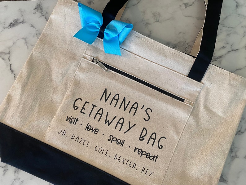 May include: A white canvas tote bag with black handles and a black zippered pocket. The bag has the text "NANA'S GETAWAY BAG visit love spoil repeat JD, HAZEL, COLE, DEXTER, REY" printed on it.