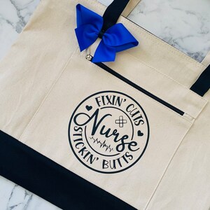 May include: A white canvas tote bag with black handles and a black zippered pocket. The bag has a black circle design with the text "Fixin' Cuts Nurse Stickin' Butts" and a heart and a plus sign.