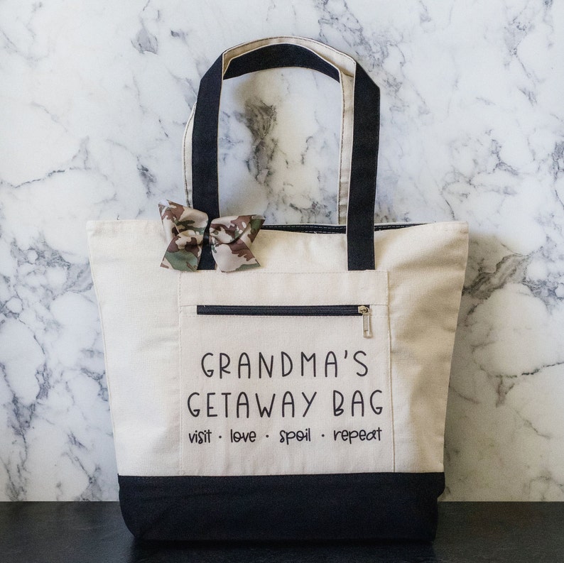 May include: A white canvas tote bag with black handles and a black bottom. The bag has a zippered pocket on the front and the text "Grandma's Getaway Bag" printed in black. Below the text is the phrase "visit. love. spoil. repeat" also printed in black.