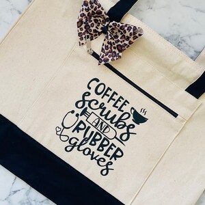 May include: A white canvas tote bag with black straps and a black bottom. The bag has a front pocket with the text "Coffee, Scrubs and Rubber Gloves" printed in black.