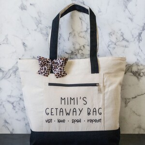 May include: A white canvas tote bag with black handles and a black bottom. The bag has a zippered pocket on the front and a black bow with a leopard print on the front. The text "Mimi's Getaway Bag visit. love. spoil. repeat" is printed on the front of the bag.
