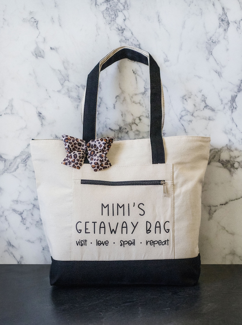 May include: A white canvas tote bag with black handles and a black bottom. The bag has a zippered pocket on the front and a black bow with a leopard print on the front. The text "Mimi's Getaway Bag visit. love. spoil. repeat" is printed on the front of the bag.