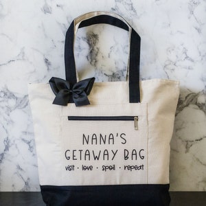 May include: A white canvas tote bag with black handles and a black zippered pocket. The bag has the text "NANA'S GETAWAY BAG visit. love. spoil. repeat" printed on it.