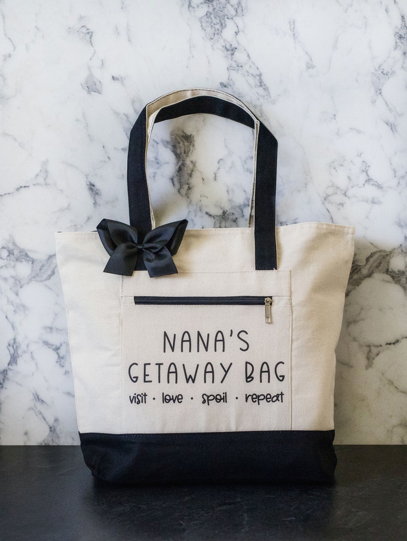 May include: A white canvas tote bag with black handles and a black zippered pocket. The bag has the text "NANA'S GETAWAY BAG visit. love. spoil. repeat" printed on it.