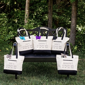 May include: Five white canvas tote bags with black handles and black bottoms. Each bag has a different name on it: Mimi's, Gigi's, Grammy's, Nana's, and Grandma's. Each bag has the text "Getaway Bag" below the name. The bags are sitting on a black bench in front of a green grassy area.