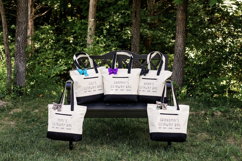 May include: Five white canvas tote bags with black handles and black bottoms. Each bag has a different name on it: Mimi's, Gigi's, Grammy's, Nana's, and Grandma's. Each bag has the text "Getaway Bag" below the name. The bags are sitting on a black bench in front of a green grassy area.