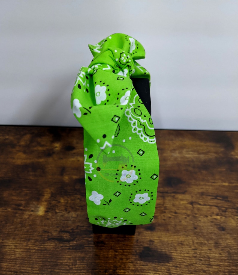 May include: Green bandana headband with a white paisley pattern. The headband is tied in a knot at the top.