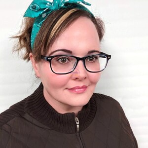 May include: A woman with brown hair wearing a teal bandana headband and black framed glasses. She is wearing a brown quilted jacket with a zipper.