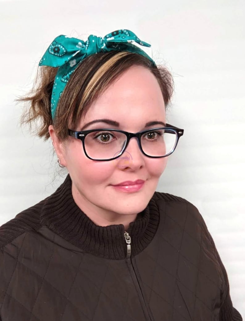 May include: A woman with brown hair wearing a teal bandana headband and black framed glasses. She is wearing a brown quilted jacket with a zipper.