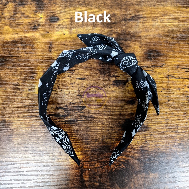 May include: Black bandana headband with a white paisley pattern. The headband is tied in a knot at the top.