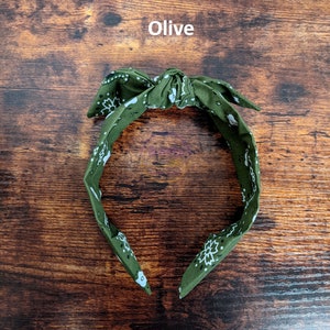May include: Olive green bandana headband with a white paisley pattern. The headband is tied in a bow at the top.